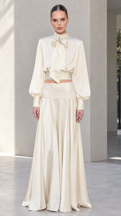 Front view of model wearing ivory satin bow tie blouse and matching pleated maxi skirt set